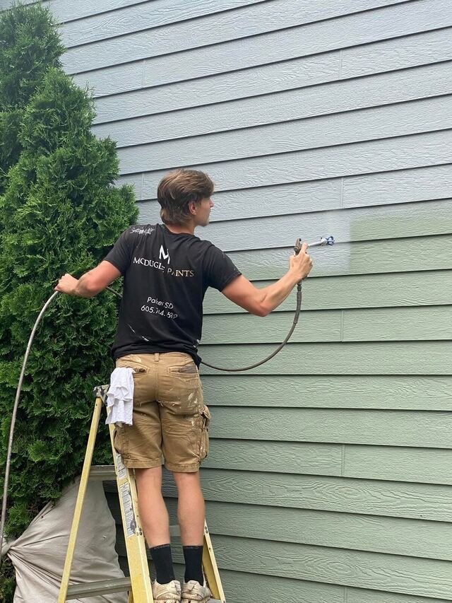 The owner of McDugle Paints on a ladder in a company shirt, spraying new paint onto lap siding